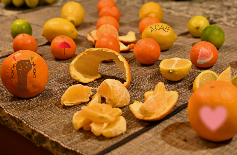 On a wooden table sits various citrus fruits, with some sliced while the orange closest to the camera is peeled with segments and skin lying on the table.  Some of the fruits have symbols/text on them (ACAB, a picture of a sliced watermelon, a  heart, a hand breaking chains).  